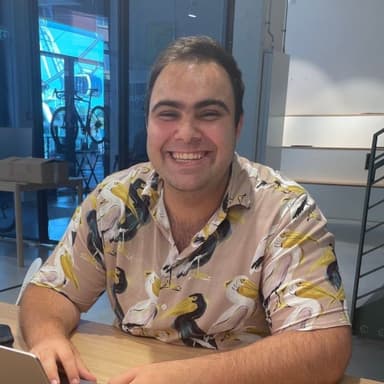 Smiling person wearing a bird-patterned shirt sits at a table with a laptop, in a modern indoor setting with glass and metal elements.